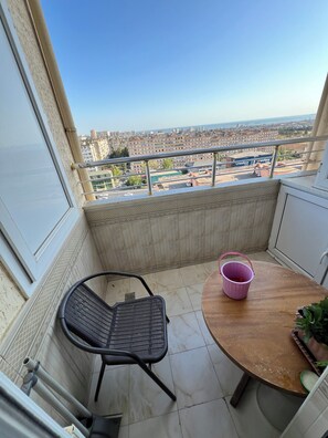A balcony with a view of a cityscape, a table with a pink bucket, and a black chair.