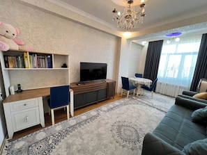 A modern living room with a flat-screen TV, a wooden desk, a sofa, and a dining area with a table and chairs.