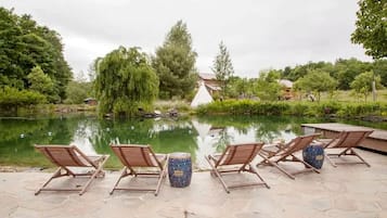 Un espacio al aire libre tranquilo con sillones de descanso y una urna decorativa, con vistas a un cuerpo de agua sereno y abundante vegetación.