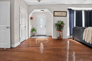 Ein Korridor mit Holzboden, einer weißen Tür, einer dekorativen Glastür, einem Blumentopf, einem gerahmten Bild und einem dunkelgrauen Sofa.