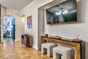 A modern living room with a large flat-screen TV mounted on the wall, a console table with a decorative bowl, and two cushioned stools. - Tropical Boho 3 bdrm house w/pool and pets are welcome. (Las Vegas)