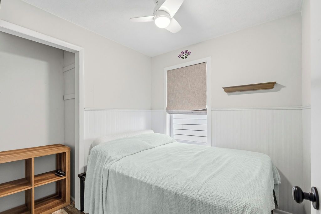 A bedroom with a bed, a wooden shelf, a ceiling fan, a window with a blind, and a small shelf on the wall.