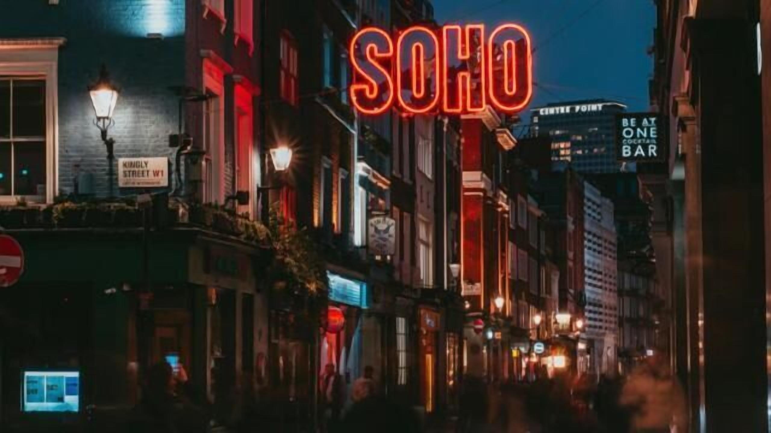 En nattbild av en gata med upplysta skyltar, inklusive en framträdande skylt med texten ”SOHO”.