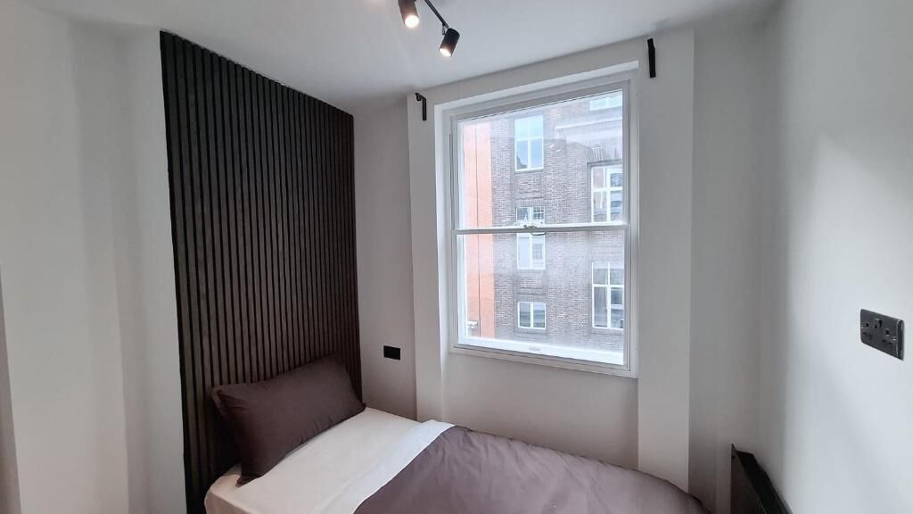Un dormitorio pequeño y moderno con una cama individual, una ventana con vistas a edificios y un techo inclinado.