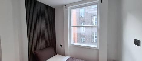 A small, modern bedroom with a single bed, a window with a view of buildings, and a sloped ceiling.