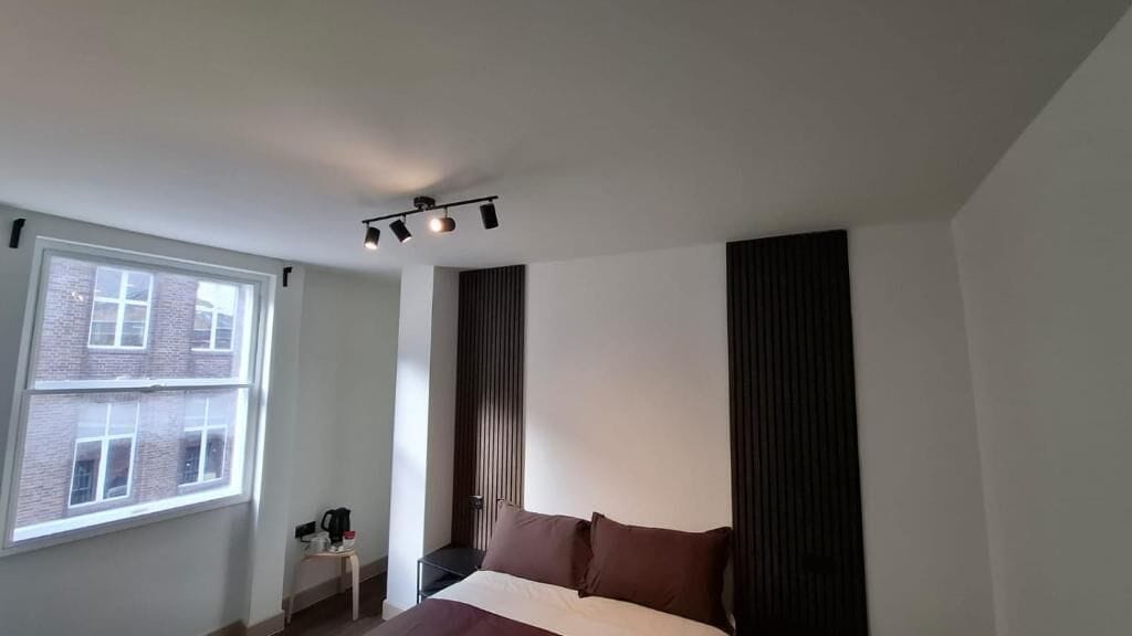 A hotel room with a bed, a window, a bedside table, and a wall-mounted light fixture.