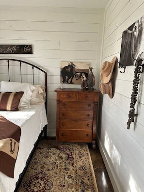 A bedroom with a bed, a wooden dresser, a wall-mounted hat rack, and a religious-themed painting. - Tejas Hills Johnson City (Johnson City)