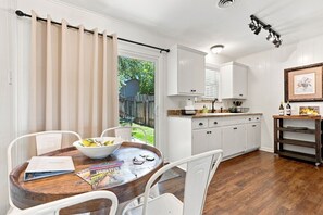 A kitchen with white cabinets, a wooden dining table with chairs, a window with curtains, and a small outdoor area visible through the window. - Tejas Hills #1 & #2 | Hill Country Pool Retreat (Fredericksburg)