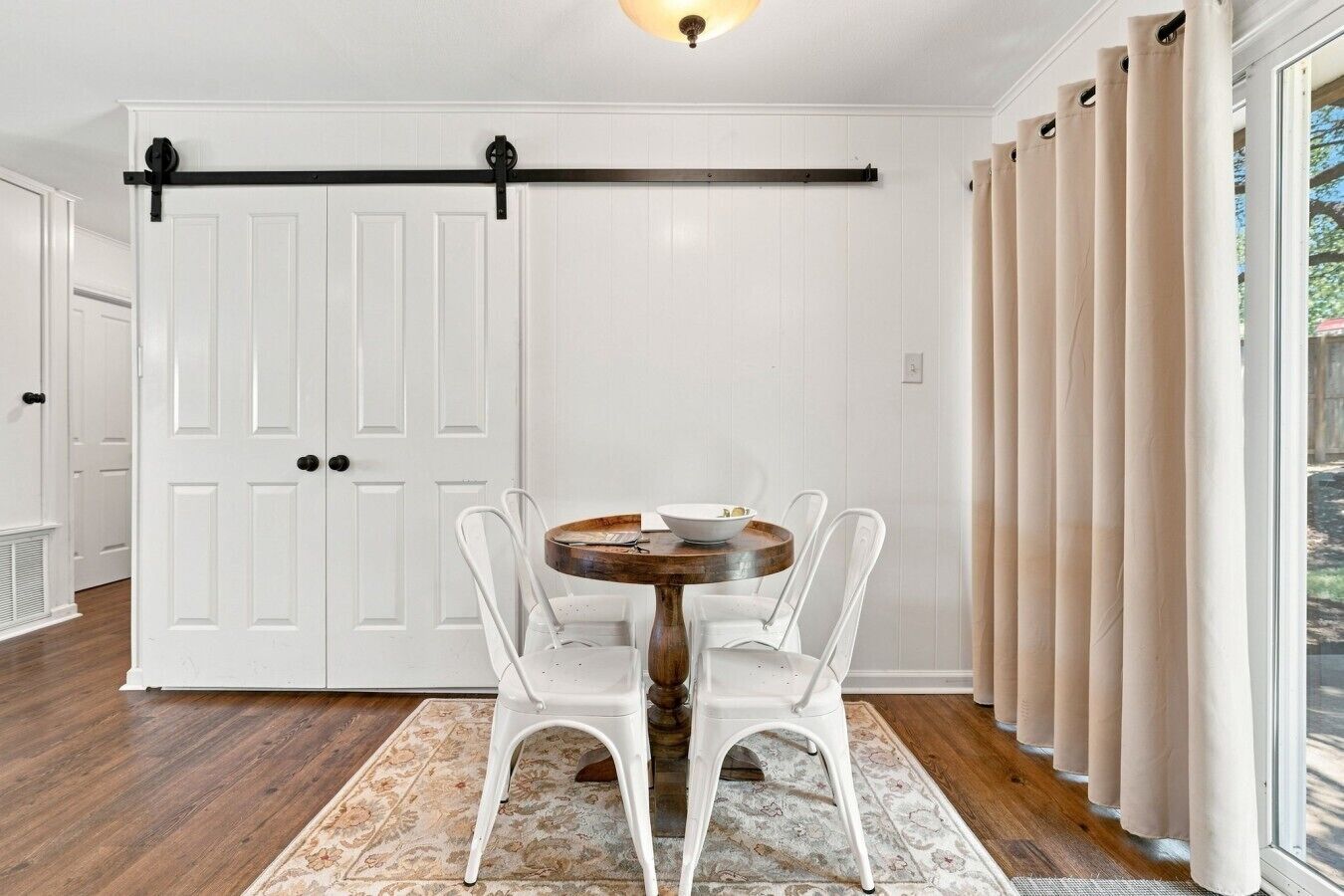 Een kamer met een witte deur, een schuifdeur in boerderijstijl, een ronde tafel met stoelen en een raam met gordijnen.