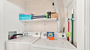 Un área de lavandería con dos lavadoras de carga frontal, un estante con ropa doblada, productos de limpieza y una caja de detergente Tide.