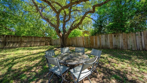 Un área de comedor al aire libre con una mesa redonda y sillas, rodeada por una valla de madera y un árbol grande.