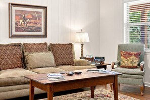 A living room with a sofa, armchair, coffee table, and a painting on the wall. - Tejas Hills Guest Haus #1 | Near Main Street (Fredericksburg)