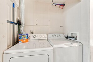 A laundry room with two front-loading washing machines, a dryer, and a box of Tide detergent. - Tejas Hills Guest Haus #1 | Near Main Street (Fredericksburg)