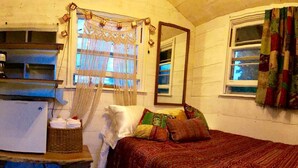 A bedroom with a bed, a mirror, a window with curtains, a refrigerator, and a basket. - Romantic Tiny Cabin Rental for a Great Vacation in the Finger Lakes Region of New York (Ithaca)
