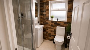 Ein Badezimmer mit Duschkabine aus Glas, einer weißen Toilette, einem Waschbecken mit Spiegel und einem Fenster mit Pflanze.