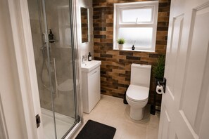 Ein Badezimmer mit Duschkabine aus Glas, einer weißen Toilette, einem Waschbecken mit Spiegel und einem Fenster mit Pflanze.