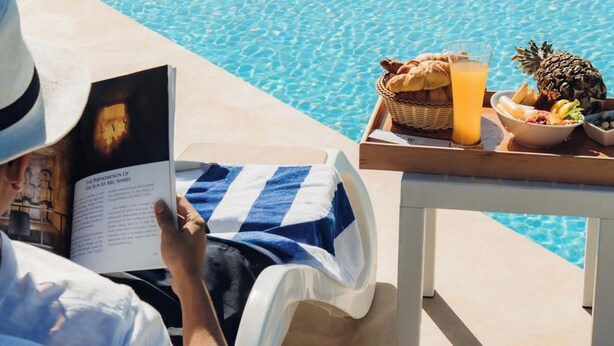 A person reading a book by a poolside with a tray of food and drinks. - Retac Abu Simbel Nefertari Hotel (Abu Simbel)