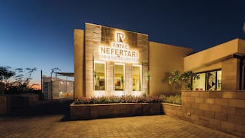 Entrada de hotel com placa indicando 'Rabat Neferarti Abusimbel Hotel' e um caminho pavimentado que leva à entrada.