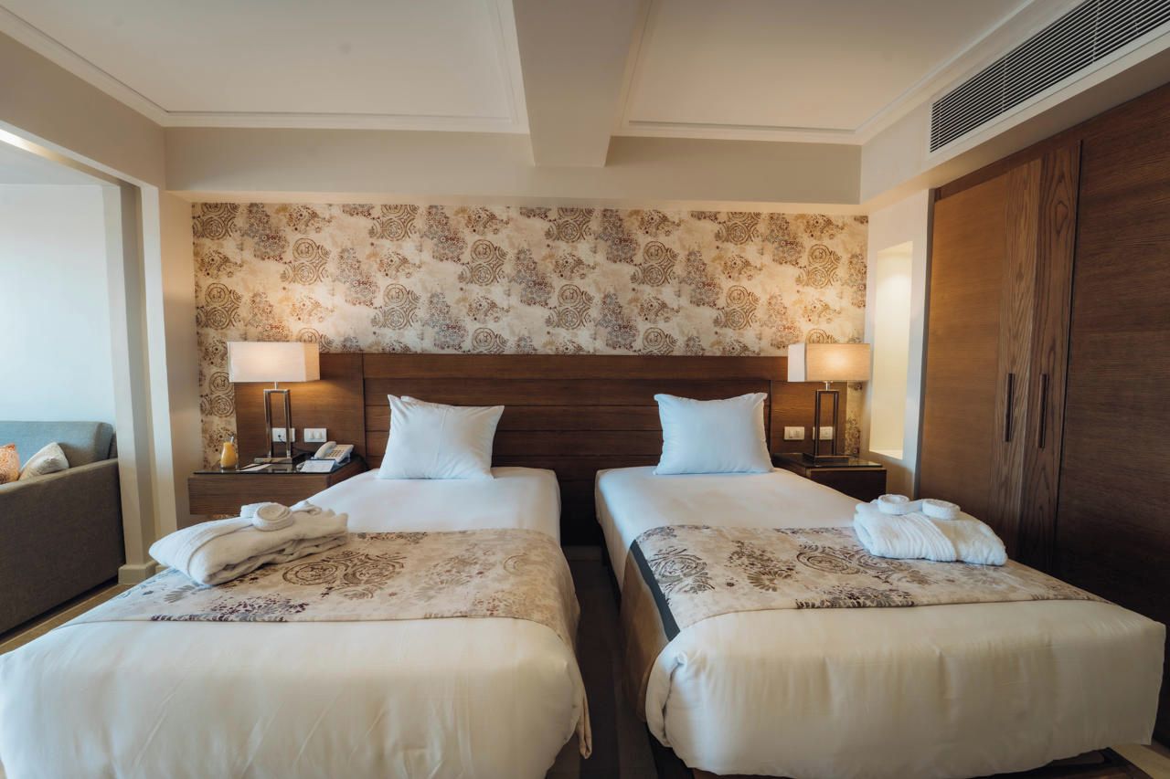 Quarto de hotel com duas camas, cabeceira de madeira, papel de parede estampado e um guarda-roupa.