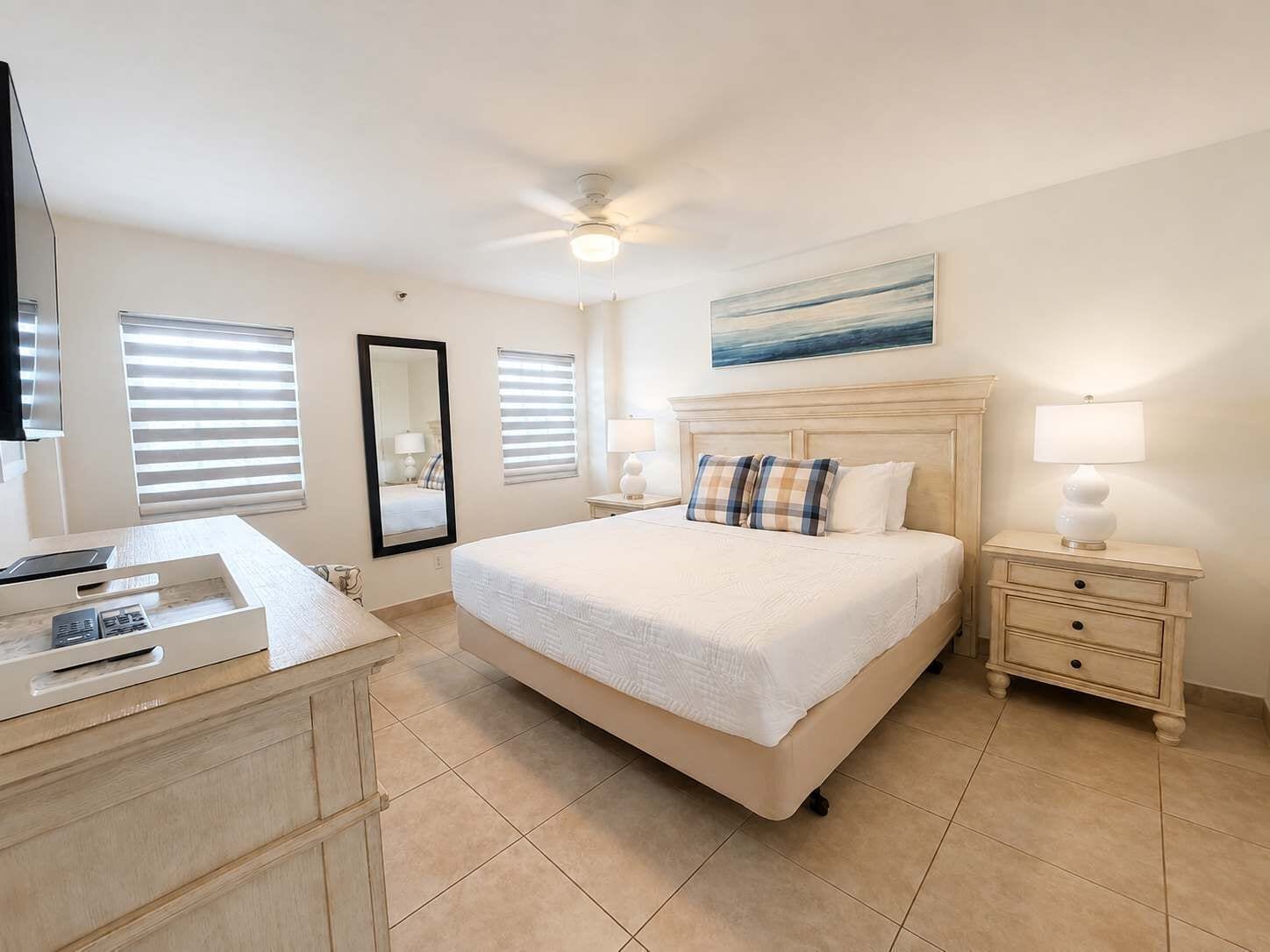 A hotel room with a bed, bedside tables, a desk with a phone, a ceiling fan, and a window with blinds.