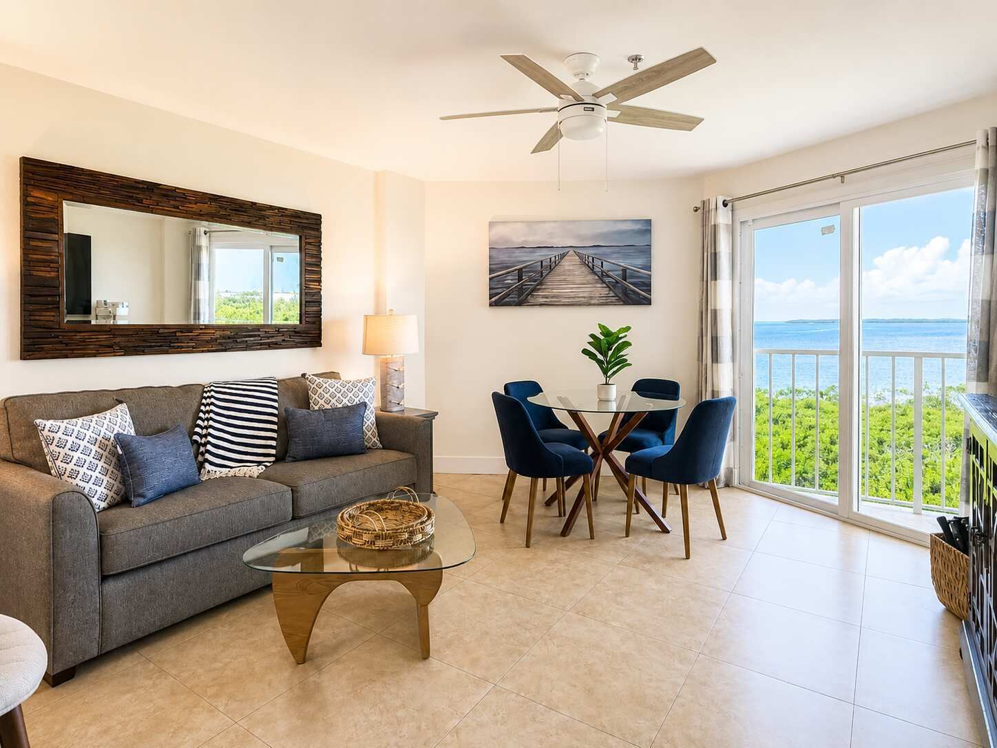 Un salon avec un canapé, une table basse et des fauteuils, une table à manger agrémentée d’une plante, un tableau représentant une jetée, et un balcon avec vue.