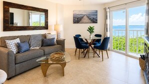 A living room with a sofa, coffee table, and chairs, a dining table with a plant, a painting of a pier, and a balcony with a view. - Key Largo Resort 2bd/2bth Ocean Views (Tavernier)