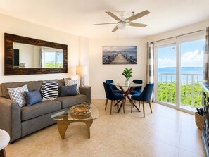 Ein Wohnzimmer mit einer Couch, einem Couchtisch und Sesseln, einem Esstisch mit einer Pflanze, einem Bild von einem Pier und einem Balkon mit Blick.
