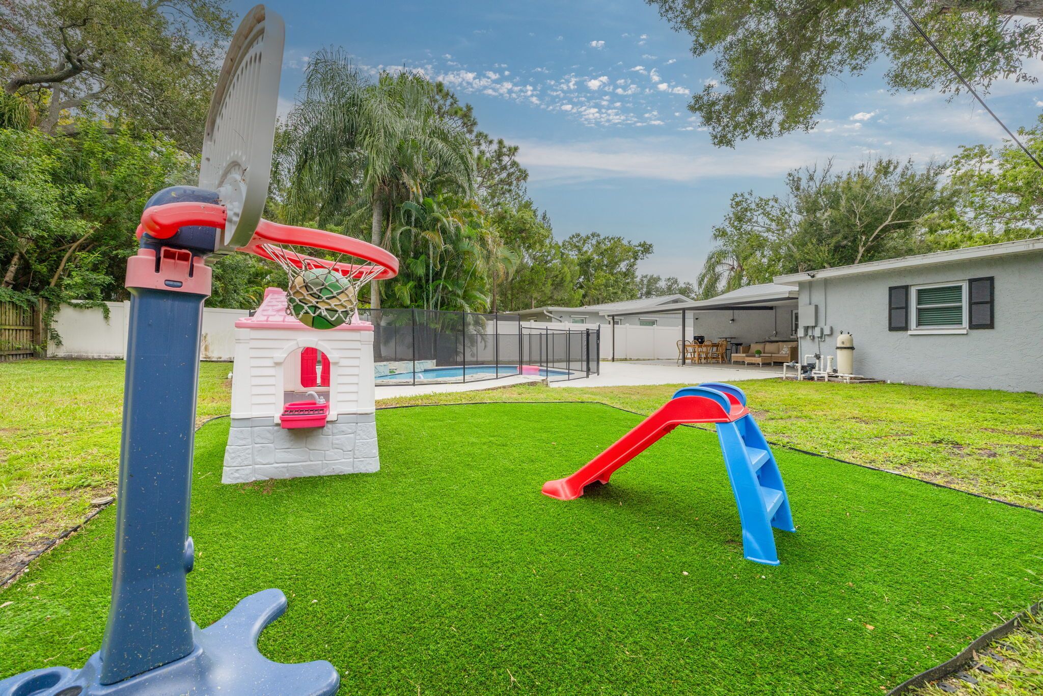 A backyard with artificial grass, a basketball hoop, a slide, and a small pool.
