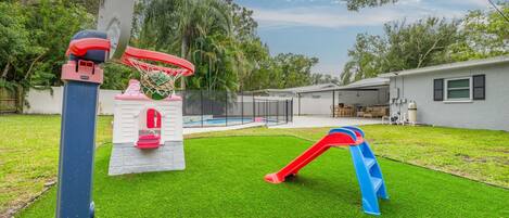 A backyard with artificial grass, a basketball hoop, a slide, and a small pool.