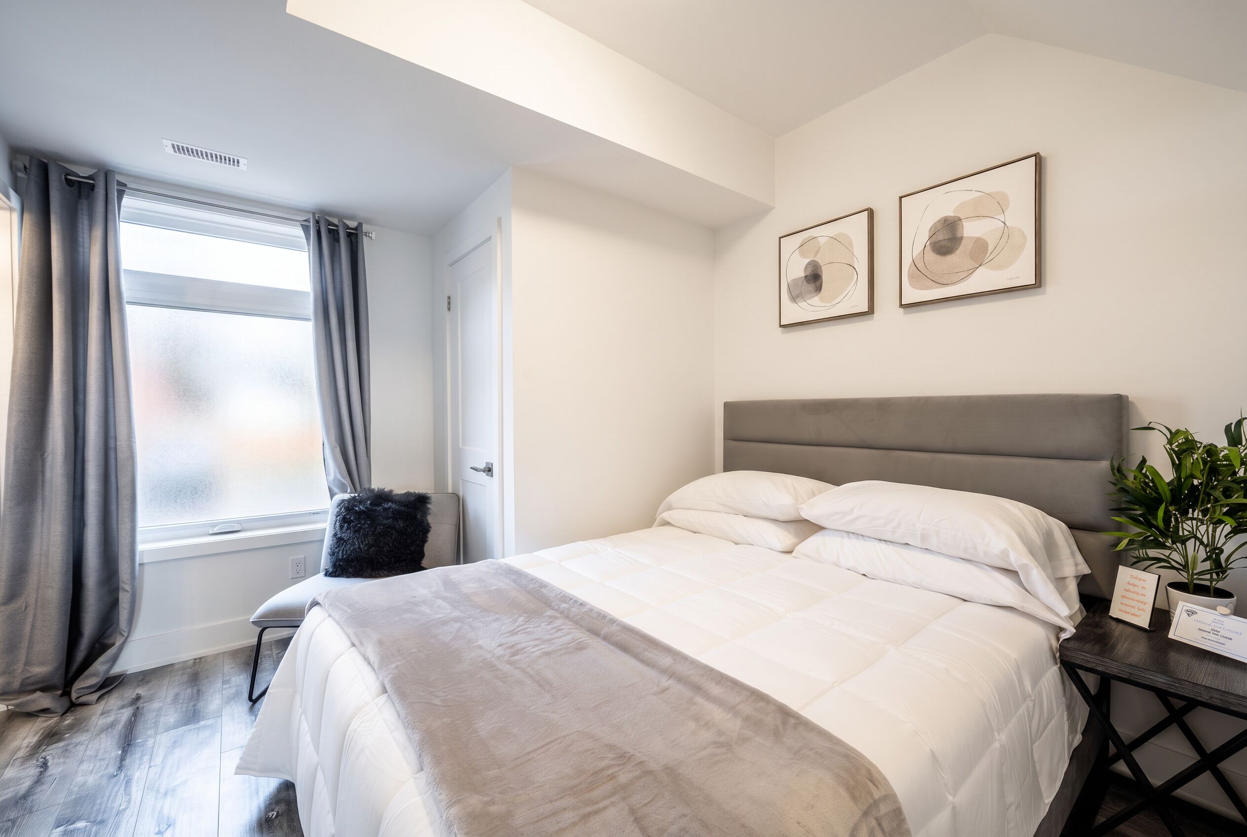 Een slaapkamer met een bed, een raam met gordijnen, een nachtkastje met een plant en twee kunstwerken aan de muur.