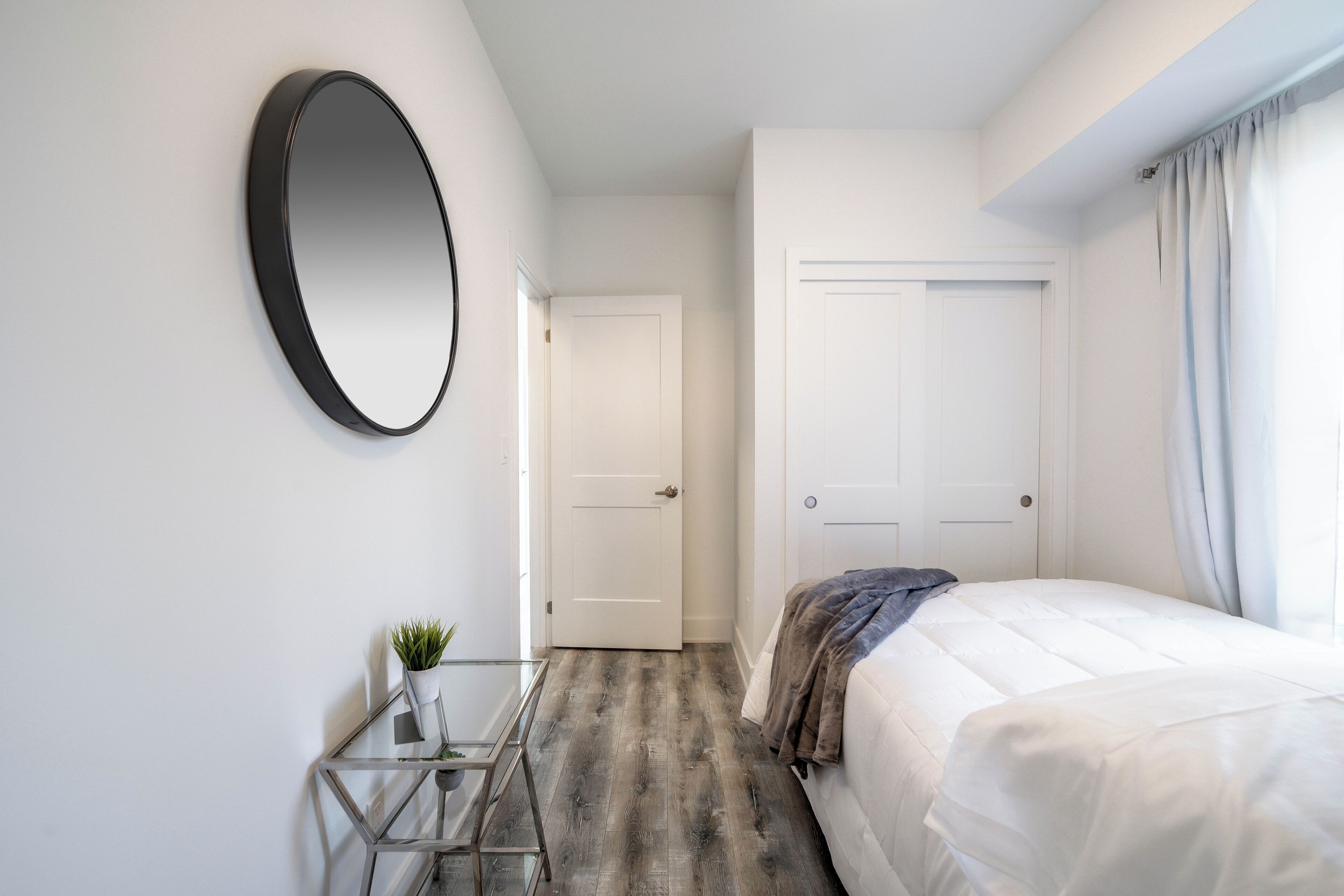 A bedroom with a bed, a round mirror, a small table with a plant, and a door leading to another room.