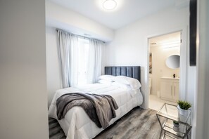 A modern bedroom with a bed, a glass table, and a bathroom visible through an open door. - Cozy 2BR Escape With Parking, 5 Mins to The Falls (Niagara Falls)