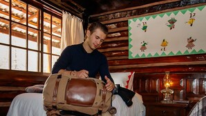 Una persona sentada en una cama con un bolso marrón grande, paredes de madera, una ventana con cortinas y un muro con un diseño verde en zigzag.