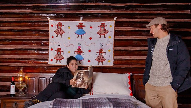 Una habitación con una cama, un muro de madera, un tapiz con figuras, una botella y un vaso.