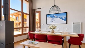En matplats med ett träbord för fyra personer, röda stolar, en bänk, ett stort fönster med utsikt över byggnader, en inramad tavla på väggen och en hängande lampa.