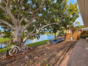 Área de pátio com mesa e cadeiras de madeira, uma bicicleta e um conjunto de churrasqueira próximo a um corpo d'água.