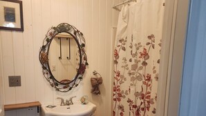 Een badkamer met een witte wastafel, een ronde spiegel met bloemenversiering, een douchegordijn met bloemenprint en een rode badmat.