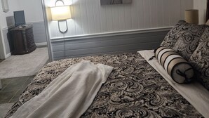 Een slaapkamer met een bed, een bedlampje, een nachtkastje, een kapstok, een spiegel en een schilderij aan de muur.
