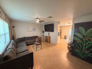 Ein Wohnzimmer mit einem Ecksofa, einem Couchtisch, einem Deckenventilator, einem Flachbildfernseher und einer Küchenzeile im Hintergrund.
