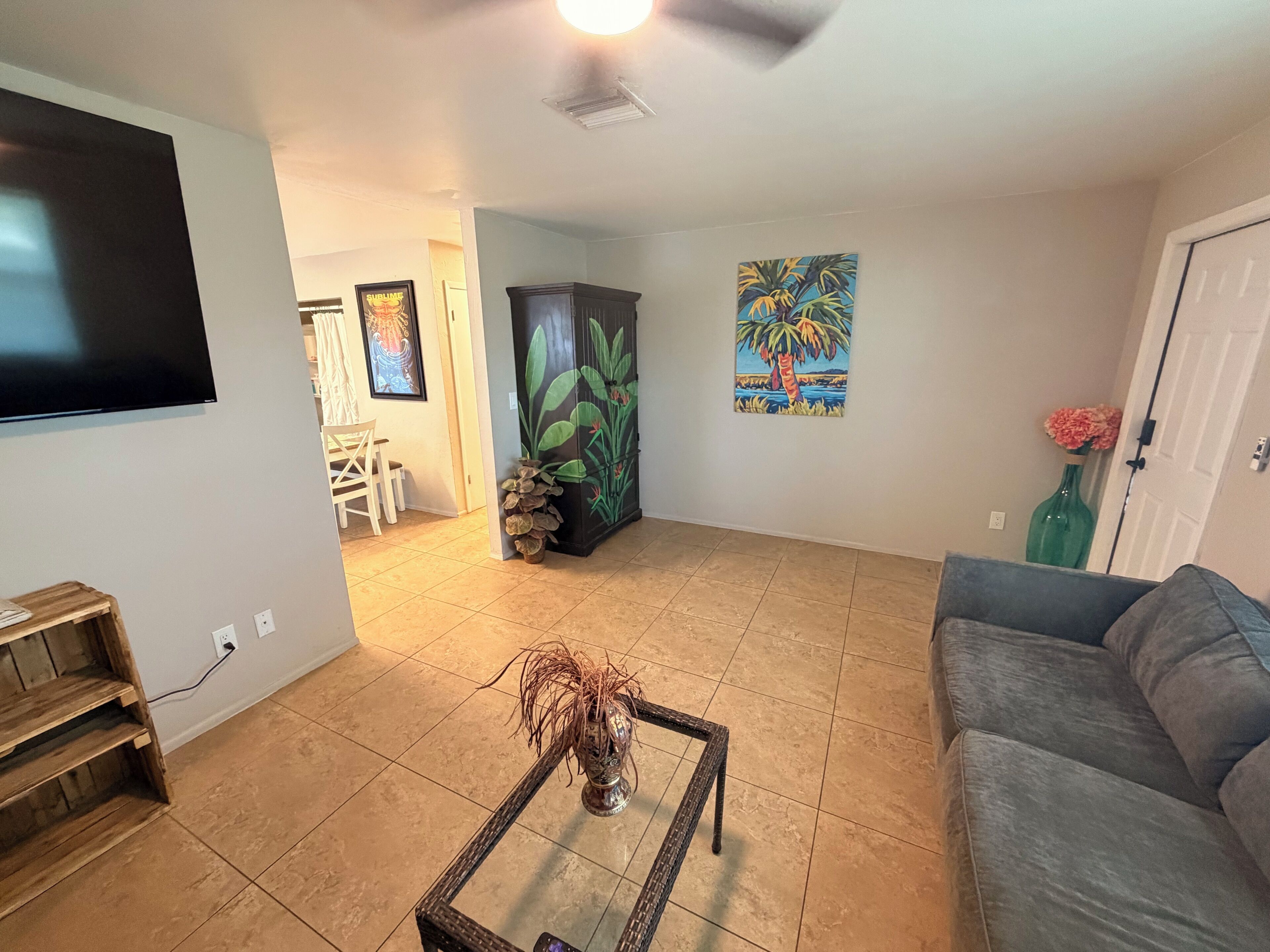 A living room with a television, a sofa, a coffee table with a decorative vase, a painting of a palm tree, and a door.