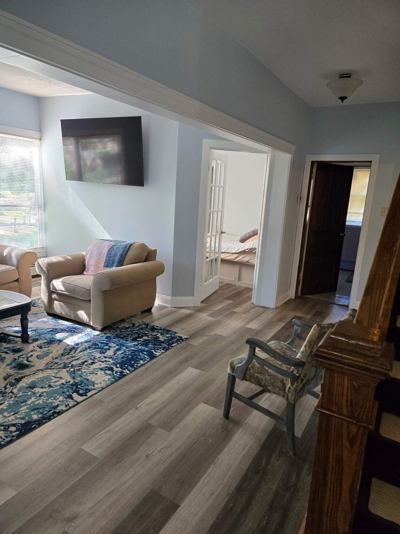 A living room with a sofa, a coffee table, and a patterned rug. There is a door leading to a bedroom with a bed.