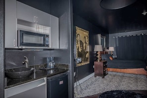 A compact room with a kitchenette including a microwave, coffee maker, and sink. There is a bed with a zebra-patterned rug and two bedside lamps.