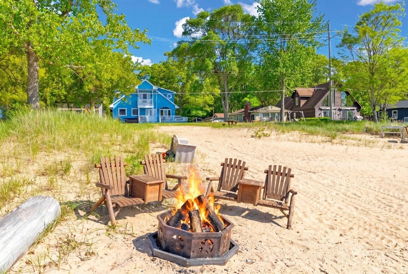 A beach scene with a fire pit surrounded by wooden chairs, a blue building in the background, and a sandy beach.