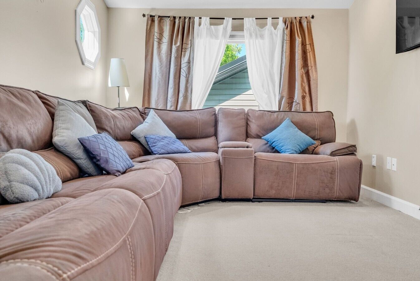 A corner sectional sofa with multiple cushions, a lamp, and a view of the outdoors through a window with curtains.