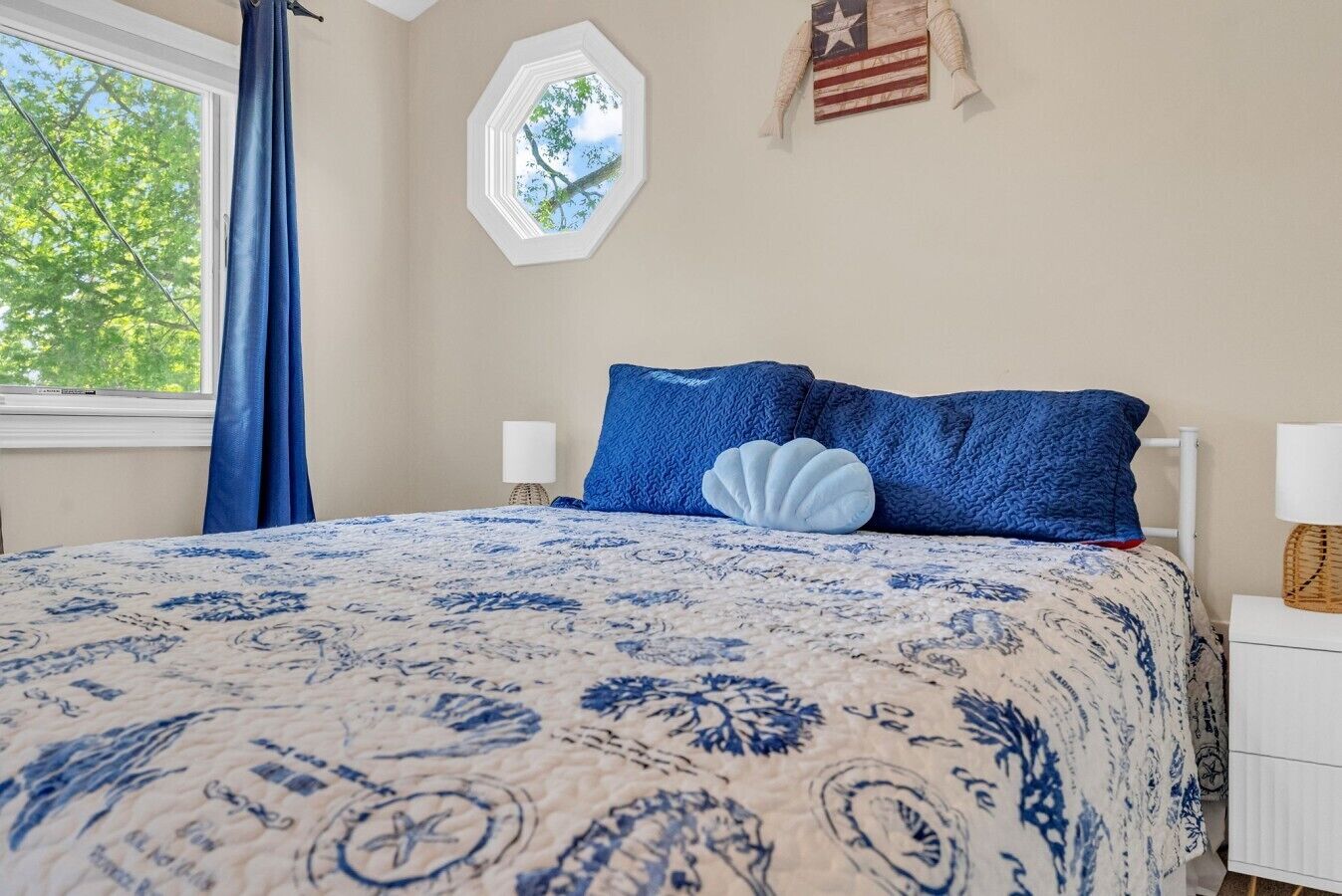 A bedroom with a bed, a window with curtains, a wall clock, and a small window with a view of trees.