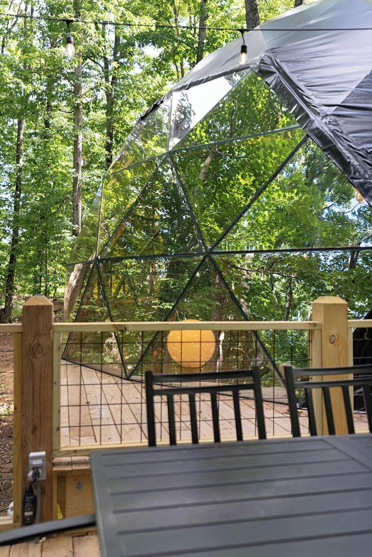 A covered outdoor seating area with a table and chairs, surrounded by a wooden fence and trees.