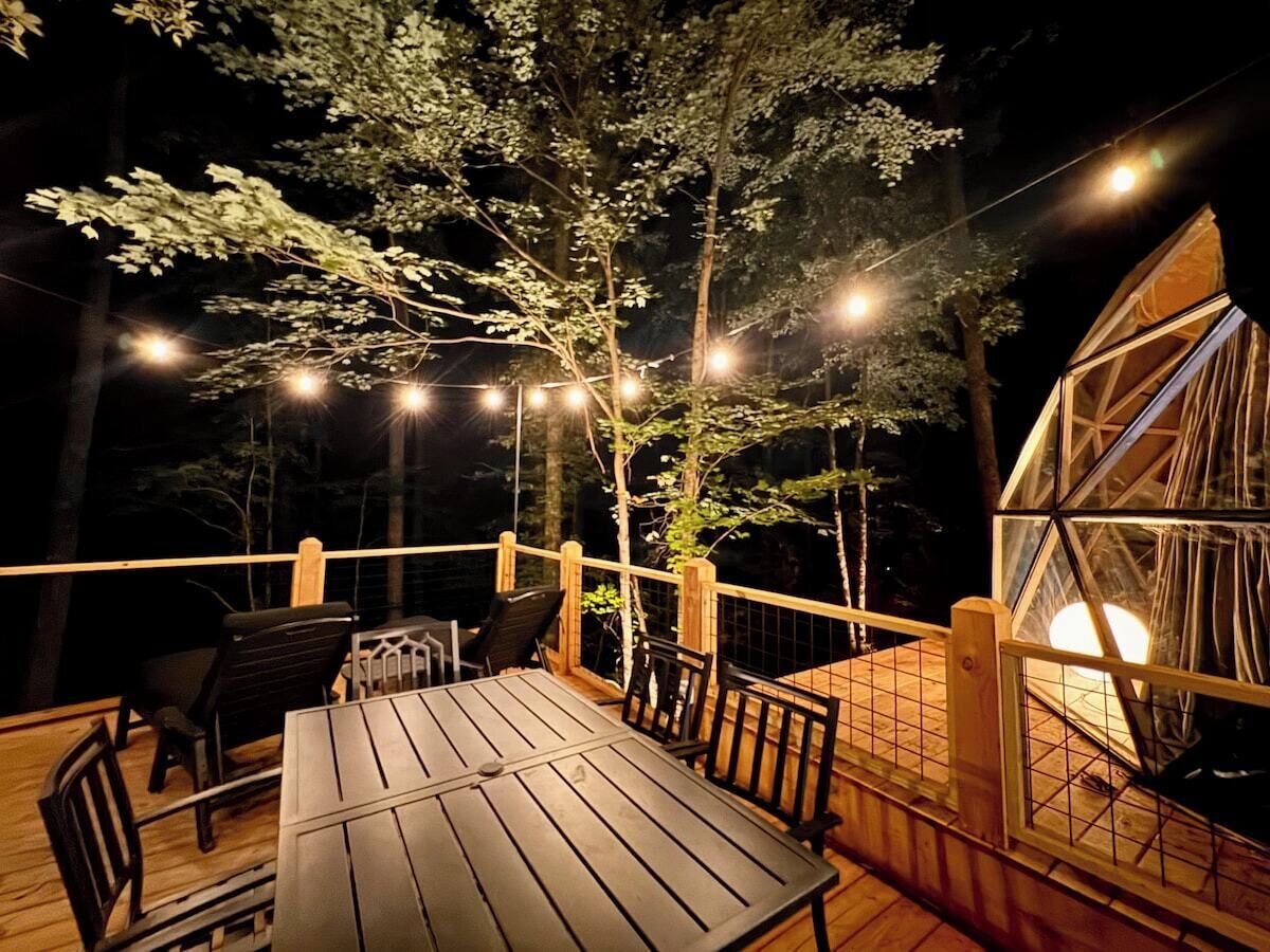 Una terraza de madera con una mesa y sillas, rodeada de árboles y luces colgantes.