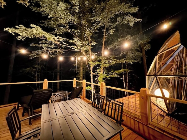 A wooden deck with a table and chairs, surrounded by trees and string lights.