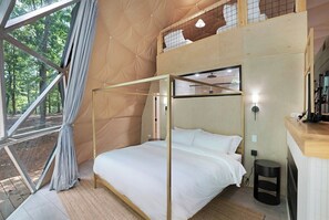 Una habitación con cama, una mesita, ventana con cortinas y vista a árboles.