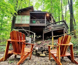 Une cabane avec une terrasse, deux fauteuils Adirondack et un foyer en pierre.